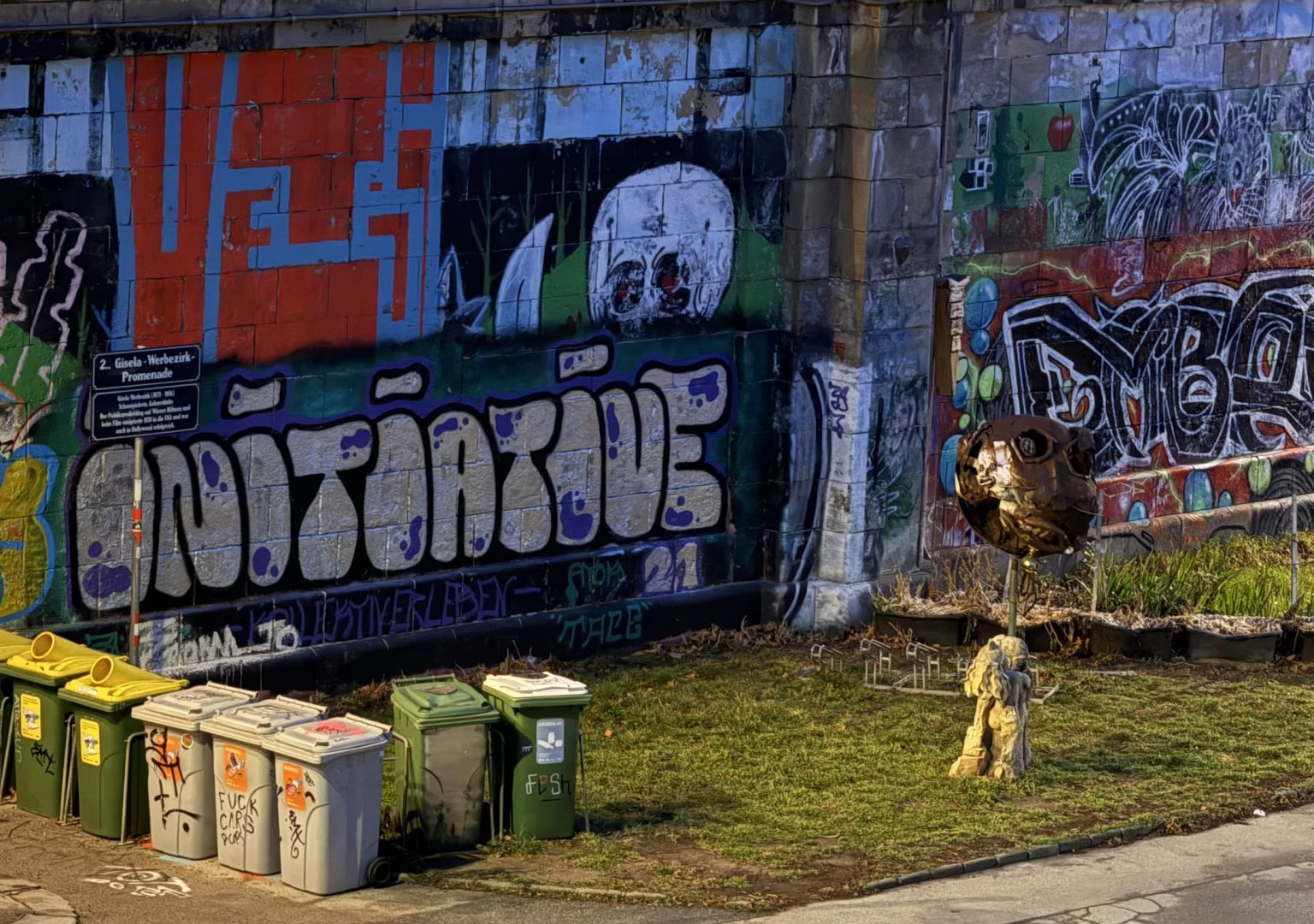 Eine mit Graffiti bedeckte Mauer mit verschiedenen bunten Schriftzügen und Motiven, darunter ein stilisierter Totenkopf und das Wort “Initiative” in großen weißen Buchstaben. Davor stehen mehrere Mülltonnen in verschiedenen Farben, einige mit Aufklebern oder Graffiti versehen. Rechts im Bild befindet sich eine Skulptur aus Metall und Stein, die einen Kopf darzustellen scheint. Die Szenerie befindet sich an einer Grünfläche mit einem kleinen Schild, das auf die “Gisela-Werbezirk-Promenade” hinweist.