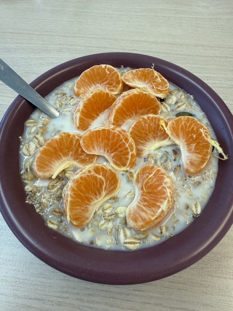 Ansicht von oben. Schale mit Müsli in Milch. Darauf sind Mandarinenstücke drapiert. Ein Löfel steckt im Müsli.